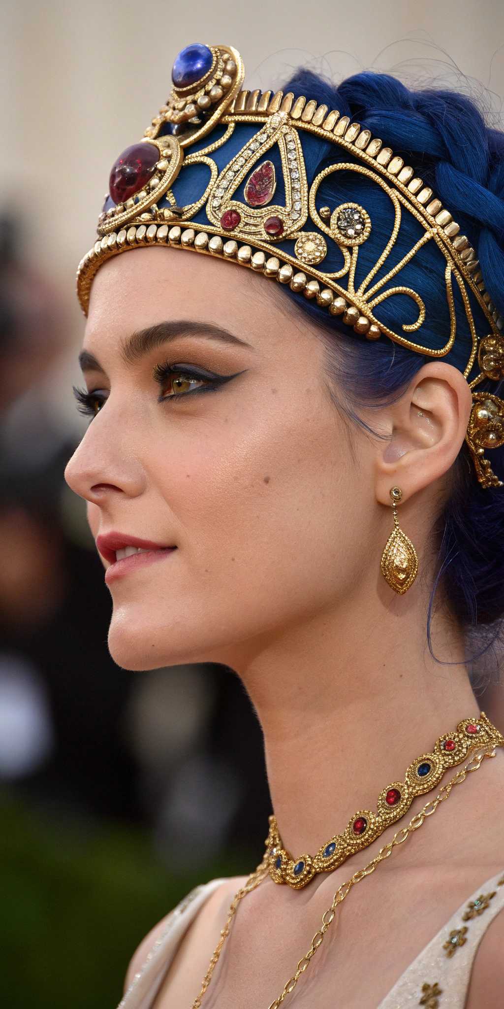 This image is a digital artwork that captures a closeup portrait of a woman. The art style is reminiscent of Art Nouveau or Art Deco, with its intricate details and ornamental elements. The medium appears to be digital painting, given the smooth blending of colors and the lack of texture that might be present in a traditional painting.The colors in the image are rich and vibrant. The womans hair is a deep, royal blue, which stands out against the golden tones of her accessories. The golden headpiece she wears is adorned with a large, ornate blue flower with a golden center, which adds a pop of color to the composition. The circular background behind her is a gradient of gold, with a complex pattern that suggests a celestial or mechanical theme.The womans makeup is done in a style that echoes the opulence of the Art Deco era, with bold, winged eyeliner and a matte red lipstick that contrasts sharply with her pale skin. Her eyes are accentuated with dark, defined eyebrows and subtle eyeshadow, giving her a piercing gaze.She is wearing a necklace with multiple strands of pearls and a pendant that matches the blue of the flower in her hair. The necklace cascades down her collarbone, adding to the luxurious feel of the image.The overall mood of the image is one of elegance and opulence, with a touch of mystery. The womans expression is enigmatic, and the intricate details of her attire and the background suggest a world of wealth and sophistication.
