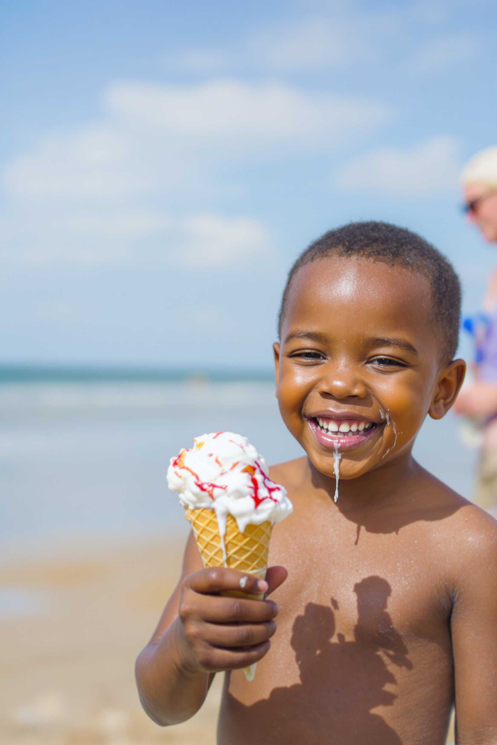 A joyful African American toddler boy with a big smile on his face, holding a dripping ice cream cone on a sunny day at the beach.

