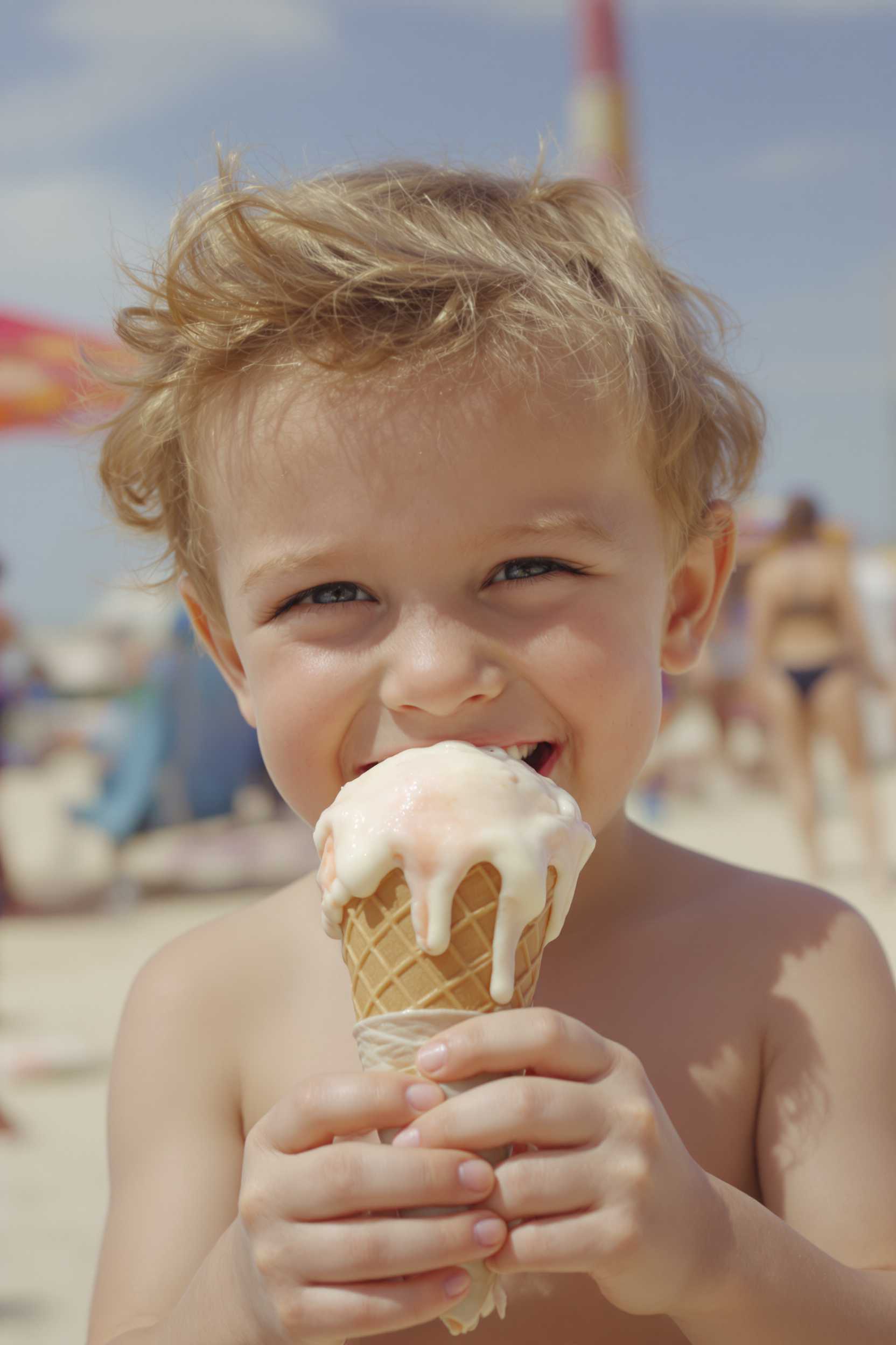 A joyful 1950's toddler boy with a big smile on his face, holding a dripping ice cream cone on a sunny day at the beach.

