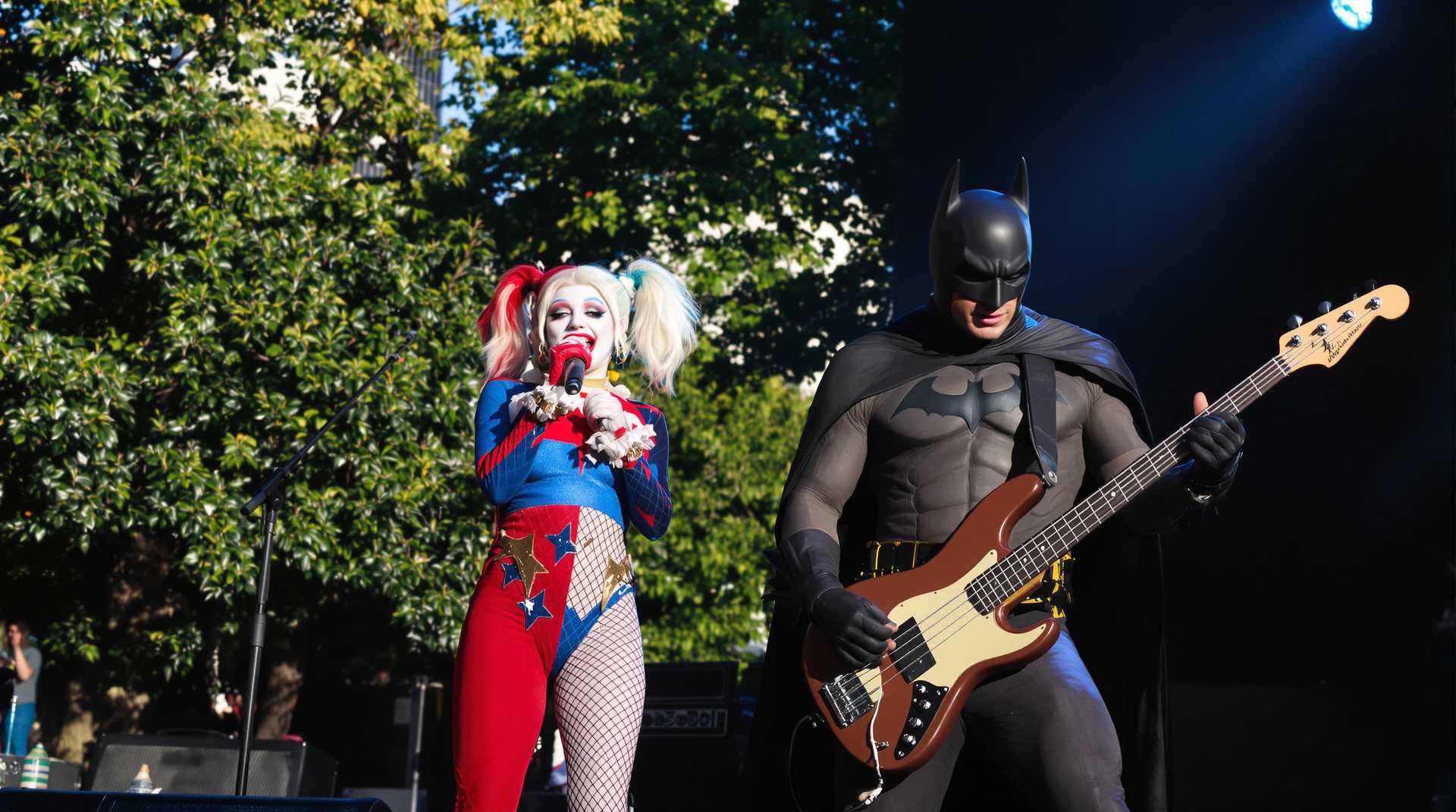 In a vibrant urban park setting, with the city skyline visible in the distance, the scene captures Harley Quinn standing at center stage, holding a microphone passionately as she sings. She is dressed in her iconic suit, with vibrant hues of red  and blue, catching the glint of the late afternoon sun. To her right, Batman rocks out on an brown 'Alembic' Stanley Clarke model electric bass guitar, his cape flowing dramatically as a spotlight emphasizes his formidable presence.