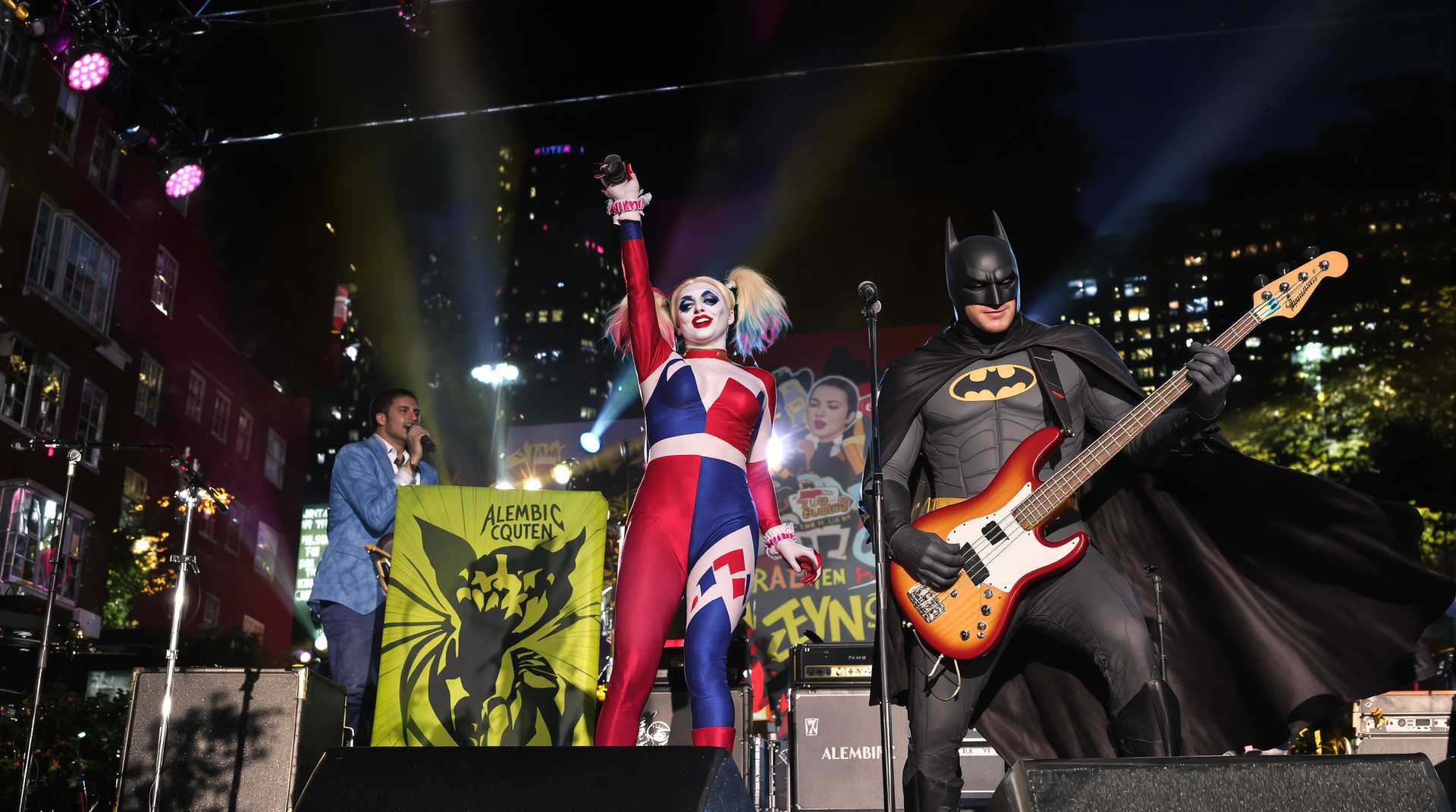 In a vibrant urban park setting, with the city skyline visible in the distance, the scene captures Harley Quinn standing at center stage, holding a microphone passionately as she sings. She is dressed in her iconic suit, with vibrant hues of red  and blue, catching the glint of the late afternoon sun. To her right, Batman rocks out on an 'Alembic' model electric bass guitar, his cape flowing dramatically as a spotlight emphasizes his formidable presence.