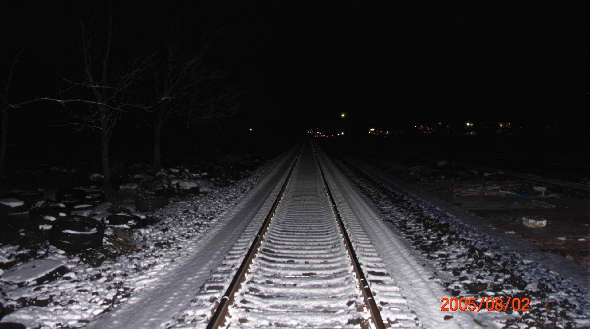 low lighting, underexposed, Snow-covered train tracks, barren landscape, leafless trees on both sides, scattered debris including tires to the left, darkness surrounds with sparse lighting from distant streetlights, wintry night, orange timestamp 2005/02/02 in the lower right corner, cold atmosphere, amateur quality, slight blur in the foreground., 
