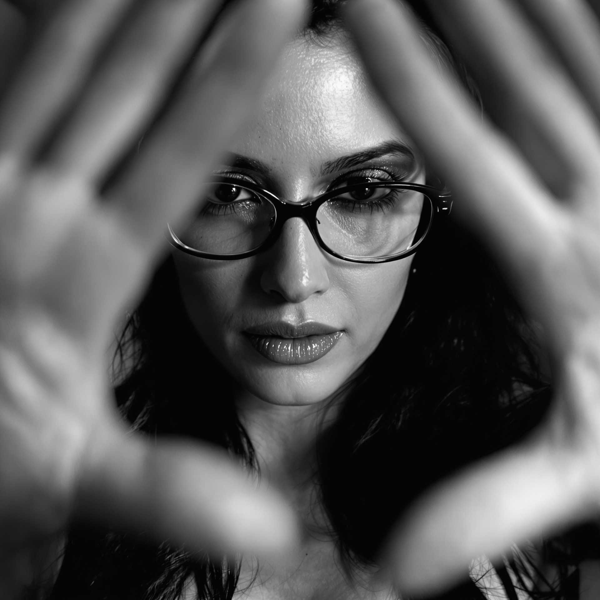 Woman. Dynamic seductive pose. Middle aged. Slender body. Glasses. Exotic. Long dark hair. Bare Chest. Deep neckline. 
Bokeh. Shallow depth of field. Cinematic photography. Sharp Image quality. High resolution. High definition.
Close up, black and white photograph featuring a person with their face framed by the blurred of her own hands. The hands framing the face are positioned in such a way that they create a sense of enclosure or intimacy around the subjects face. the hand position are away from her face, adding a dynamic element to the composition.