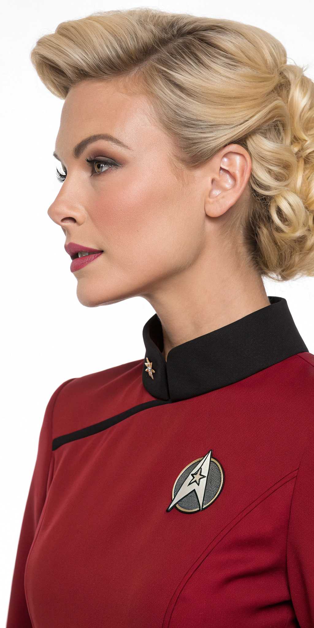 This image features a closeup of a blonde woman wearing a red Starfleet uniform from the Star Trek franchise, identifiable by the distinctive communicator badge on the left side of her large bust, chest. The uniform is a deep maroon with a black collar and a black communicator badge with a silver emblem in the center. The person is also wearing a Starfleet insignia on the left shoulder, which is a silver arrowhead with a gold outline.The hair of the person is styled in a voluminous, 1960sinspired fashion, with waves and curls cascading down the back. The hair color is a light blonde with a touch of golden highlights. The person is also wearing a pair of white earrings with a simple, elegant design.The background is blurred, but it appears to be an interior space with warm, reddish tones, possibly a corridor or room on a spaceship from the Star Trek universe. The overall art style of the image is reminiscent of a fan art or a tribute piece, paying homage to the classic Star Trek aesthetic. The medium appears to be digital, given the sharpness and clarity of the image, The colors are rich and vibrant, with a focus on the deep red of the uniform and the blonde hair, which stands out against the warmer tones of the background.