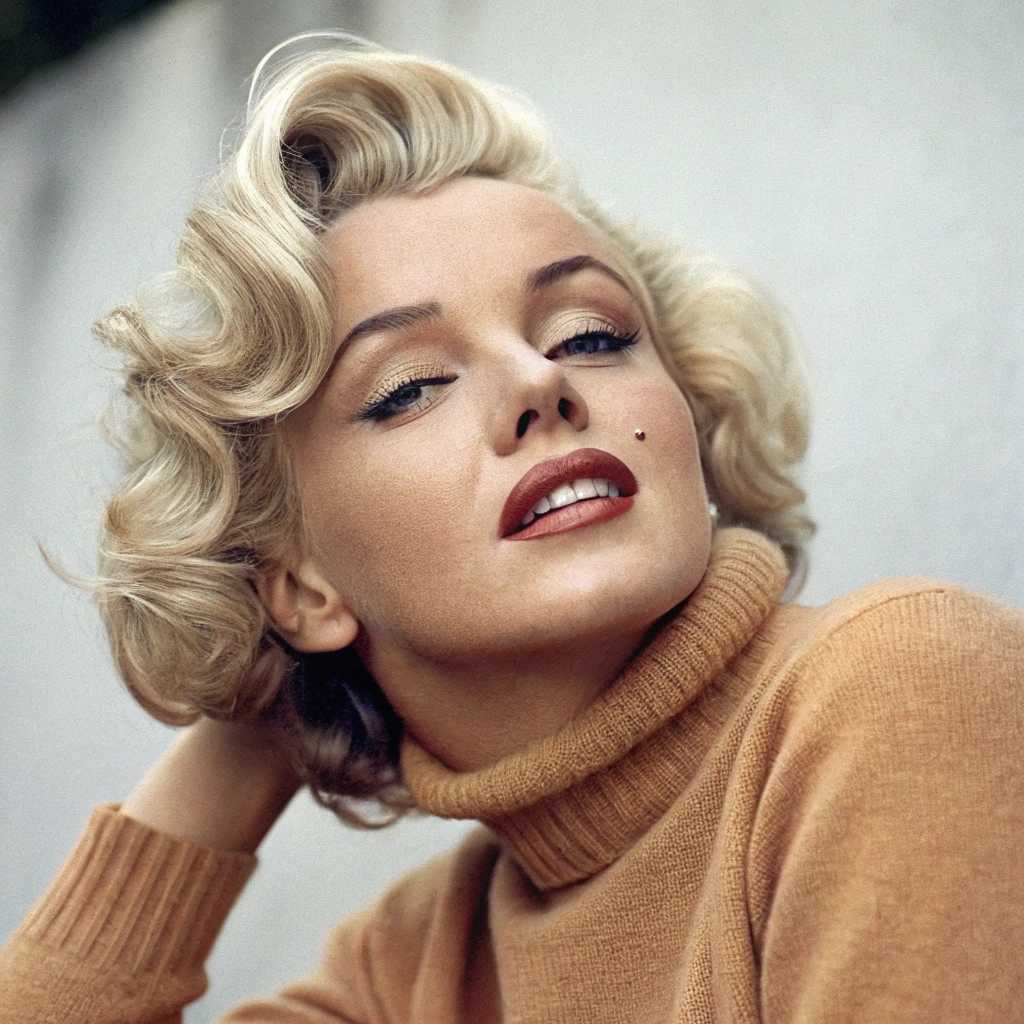 This image is a closeup portrait of "Marilyn Monroe" with a hairstyle reminiscent of the 1950s, featuring a voluminous blonde wave on the right side of the head. The hair is styled in a way that suggests a classic Hollywood glamour, with a side parting that adds to the vintage allure.The subject is wearing a high neck, warm toned sweater that appears to be made of a soft, knit fabric, which gives off a cozy and comfortable vibe. The sweaters texture is not very pronounced, but it is visible in the way the light reflects on the surface, giving it a subtle dimension.The subject is accessorized with a pair of large, circular gold earrings that have a simple yet elegant design. The earrings are attached to the subjects ears with what seems to be a single loop, and they dangle just below the earlobe, catching the light and adding a touch of opulence to the overall look.The background of the image is a neutral, muted color that fades into a soft, grayish tone, which serves to focus the viewers attention on the subjects face and hairstyle. The background is not detailed, which allows the viewer to concentrate on the subjects features and the overall composition of the image.The art style of the image is reminiscent of a classic portrait photography, with a focus on the subjects features and a timeless aesthetic. The medium appears to be a digital photograph, given the clarity and sharpness of the details.The colors in the image are warm and muted, with the blonde hair providing a pop of color against the neutral tones of the sweater and the background. The red lipstick on the subjects lips adds a bold contrast, drawing attention to the subjects expression and smile.Overall, the image exudes a sense of nostalgia and elegance, capturing a moment that feels both intimate and timeless.