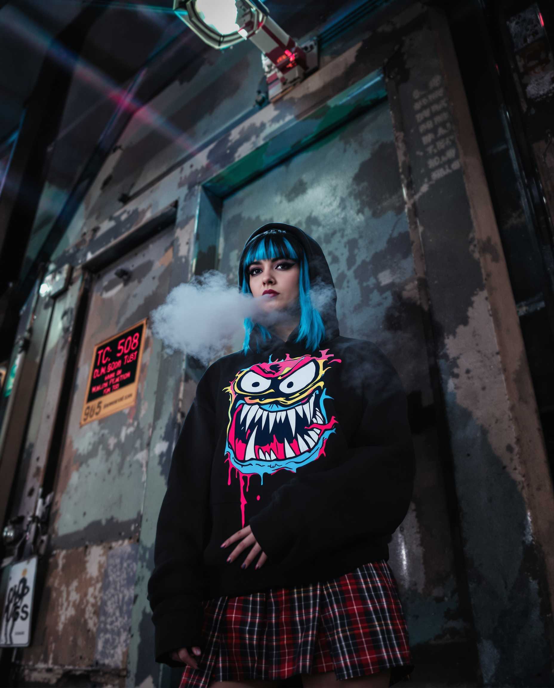 A gritty urban street portrait capturing a woman embodying emo fashion, standing against a weathered bus station. She wears a hooded sweatshirt featuring a bold, colorful cartoon monster graphic, paired with a plaid skirt. Her striking black hair is adorned with vibrant blue streaks, adding a pop of color against the subdued background. The setting is an evening scene with a dramatic cold ambiance, visible as her breath forms mist in the icy air. The lighting is emphasized by the streetlamp's glow, creating an atmospheric lens flare effect. The composition uses a low-angle, slightly tilted perspective to capture her defiant stance and the texture of the grungy environment, highlighting the edgy, raw elements of street style photography. The mood is intensified by the chilly night, enveloping the scene in a sense of solitude and introspection.