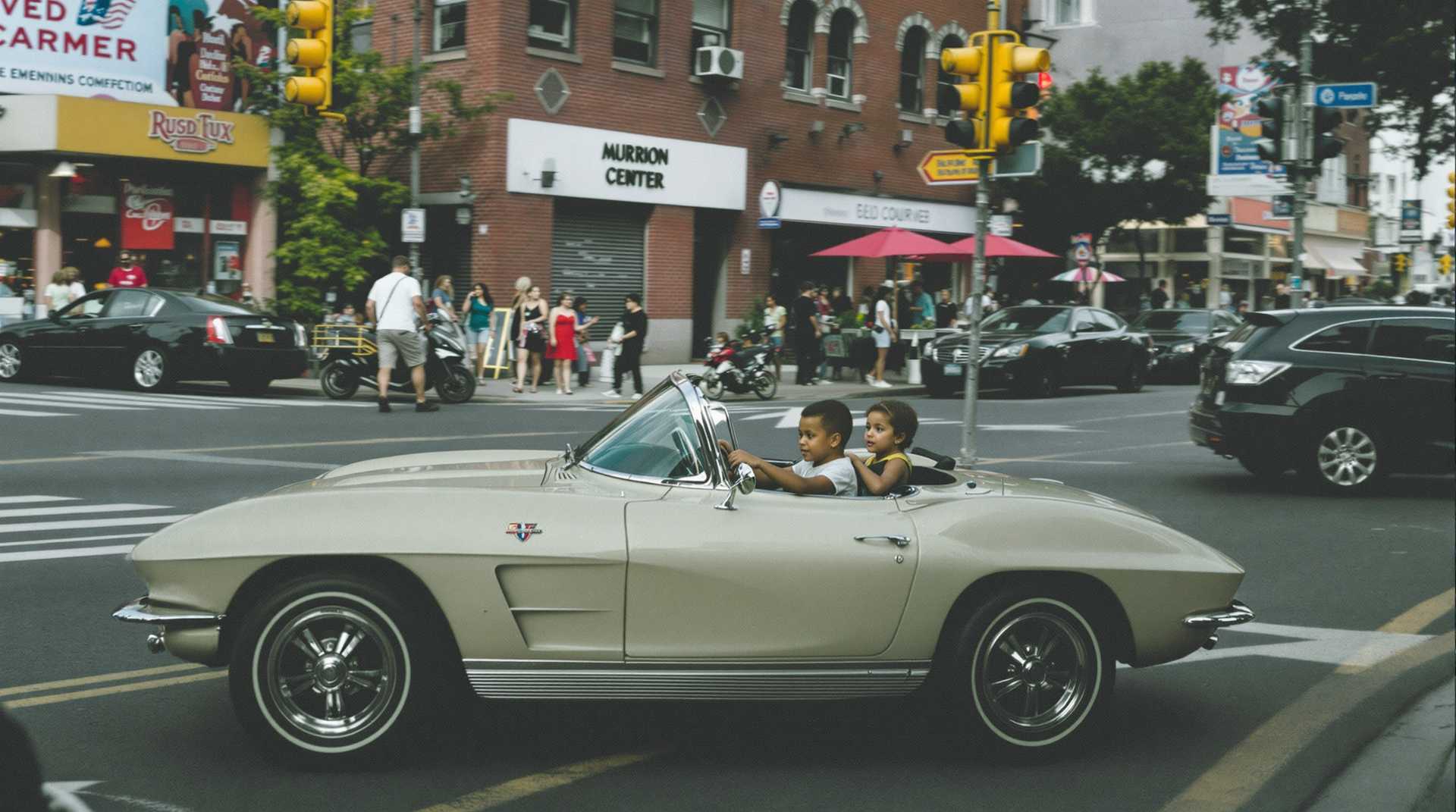 A 2 year old drives a mini 1963 corvette converible in a city street.