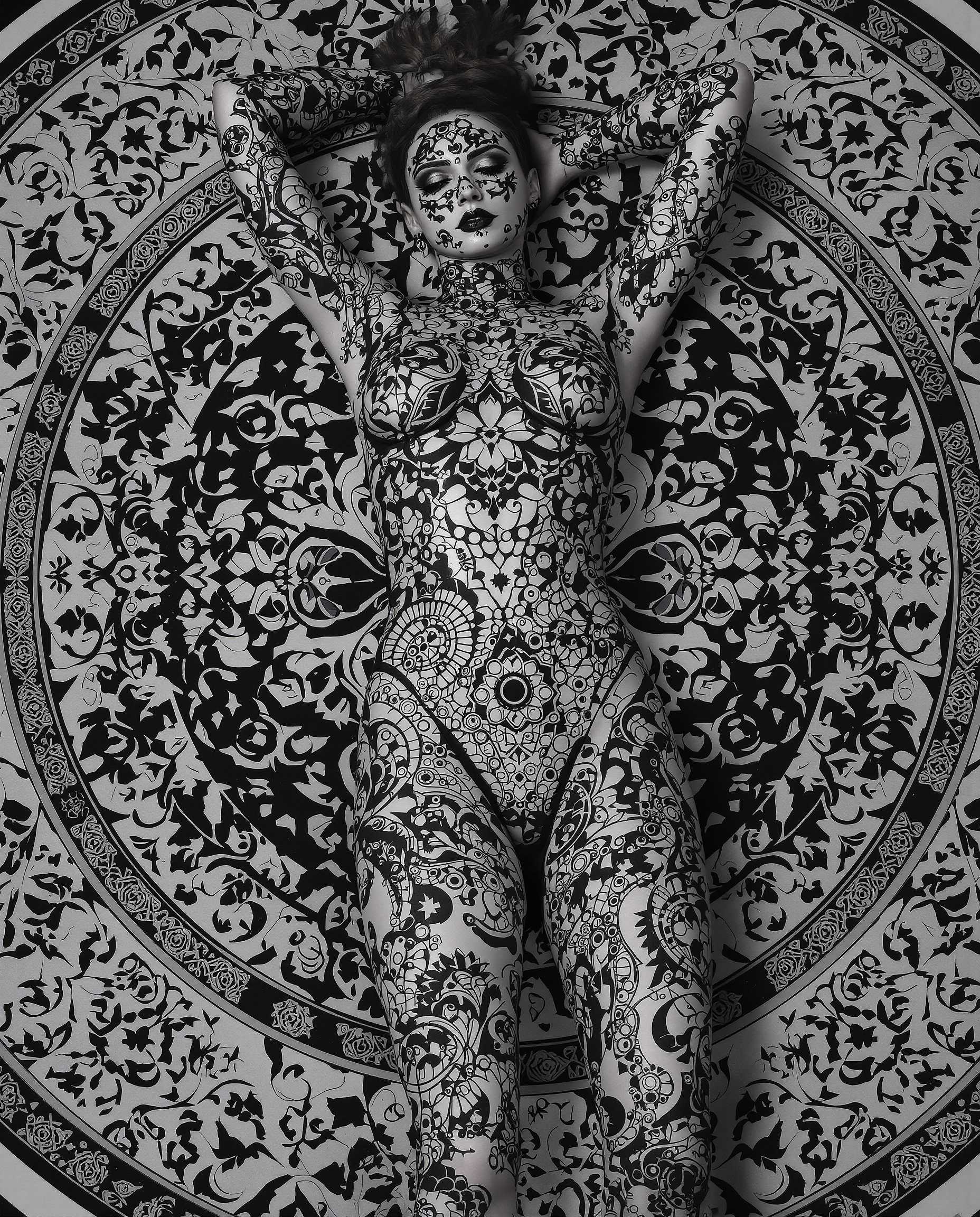 This is a realistic photo (photograph) of a female real person highly stylized and detailed digital artwork, predominantly in grayscale with a few highlights of black and white. The subject of the artwork is a figure lying on their back, with their arms resting behind their head, conveying a relaxed or contemplative pose.The figure is adorned with intricate, gothic inspired tattoos that cover their entire body. The tattoos are elaborate, with a repetitive pattern of circular motifs, floral designs, and what appear to be mechanical elements, such as gears and cogs. The tattoos are executed with precision, and the shading and highlights give them a threedimensional quality.The figure is wearing a formfitting bodysuit that complements the tattoos, with a similar gothic design. The bodysuit is made of a shiny, possibly metallic material, which reflects the light and adds to the threedimensional effect of the artwork.The figure is lying on a circular, ornate carpet or mat that has a complex, symmetrical pattern. The pattern is reminiscent of Islamic or Ottoman style tile work, with interlocking geometric shapes and floral motifs. The carpets design is monochromatic, with shades of black and white, and it provides a stark contrast to the figures tattoos and the shiny texture of the bodysuit.The overall art style of the image is realistic, with a focus on detailed line work and shading that gives the characters and objects a lifelike quality. The medium appears to be digital painting, given the smooth gradients and seamless blending of colors.The colors in the image are primarily shades of gray, with black and white accents. The grayscale palette gives the artwork a moody and atmospheric quality, while the highlights of black and white add depth and contrast.Overall, the image is a striking and visually compelling piece of digital art that combines gothic aesthetics with intricate design and shading techniques.