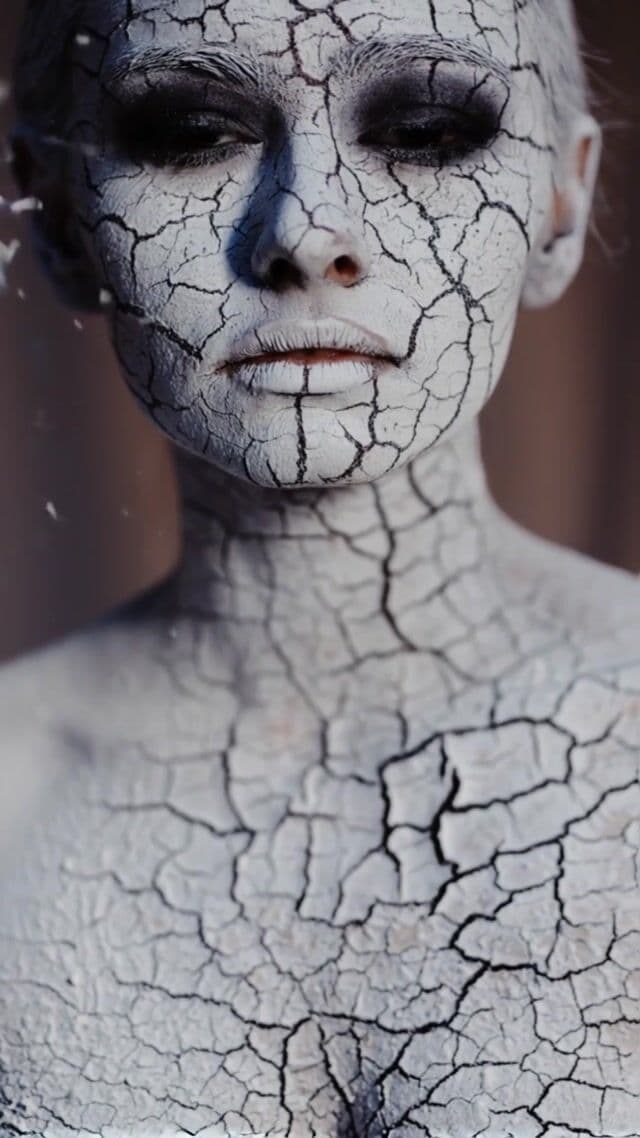 Imagine a close-up portrait of a beautiful model. Her iris is almost black, and the texture of her entire skin is white ash, with small glowing cracks visible on the surface. The wind gently blows across her face, and the cracks glow brightly, like burnt wood coals, while the wind carries away some of the ash from the surface of her skin. The background is softly blurred. Mystical, warm and mysterious atmosphere...