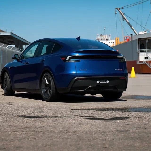 {
  "shot_composition": "A rear three-quarter view of a sleek deep blue Tesla Model Y electric crossover SUV, captured from a low ground-level angle with a wide-angle lens that emphasizes the vehicle's aerodynamic curves, black turbine-style wheels, tinted windows, and subtle badging, framed against an industrial backdrop with the car positioned centrally in the foreground.",
  "scene_setting": "An outdoor industrial port or dock area on a bright sunny day under a clear blue sky, featuring concrete ground with oil stains, white metal barriers along the sides, a covered shelter on the left, and large cranes and ships in the distant background, illuminated by natural daylight casting soft shadows on the pavement.",
  "subject_wardrobe": "",
  "motion_animation": "The deep blue Tesla Model Y starts from its parked position on the concrete surface and slowly drives off forward at a gentle pace, with its black wheels rotating smoothly and the vehicle gradually accelerating away from the camera, maintaining a realistic motion path along the straight industrial roadway while the background elements remain static.",
  "camera_movement": "The camera remains static in a fixed low-angle position, tracking the car's slow departure without panning or zooming to maintain focus on the vehicle's rear and side profile as it moves away into the distance.",
  "visual_style": "Realistic photographic style with vibrant deep blue tones on the car contrasting against the bright blue sky and neutral gray concrete, capturing a clean modern aesthetic with sharp details, natural lighting, and an open industrial mood."
}