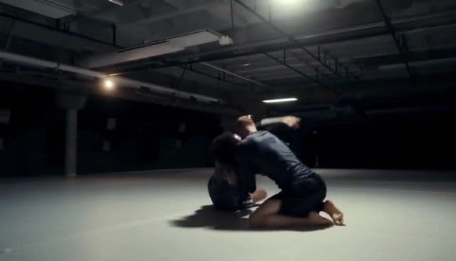 Cinematic wide-angle video of two athletes sparring in No-Gi Brazilian Jiu-Jitsu, both wearing dark rashguards. Brutalist basement gym with a pristine white mat. The camera circles slowly at a low angle, capturing their intense ground exchanges. Sweat glistens under low, moody lighting as dramatic shadows stretch across the mat. Subtle motion blur emphasizes speed, scrambles, and transitions. Highlight athletic physiques, raw determination in their expressions, and the authentic atmosphere of a real fight gym. Bold, cinematic composition with a gritty, realistic sports aesthetic.