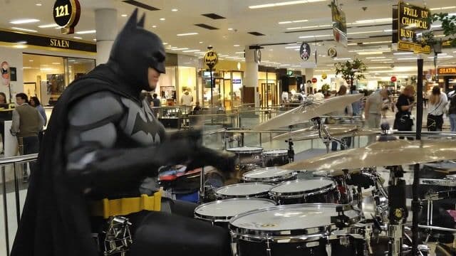 In a bustling daytime shopping mall atrium filled with multi-level walkways, storefronts, and a golden statue, Batman in his iconic black cowled suit with gray bat emblem sits at a full drum kit of black snare, red toms, and chrome cymbals, energetically delivering a "jazz fusion" drum solo with rapid stickwork, foot pedal drives, and improvisational flair. The camera starts with a medium-wide 35mm shot on Sony A7S III, slowly dollying in to capture his focused intensity and rhythmic head nods amid the murmuring crowd, under bright natural window light mixed with fluorescents. Subtle film grain enhances the lively, superhero-meets-music atmosphere as ambient mall sounds echo the dynamic beats.