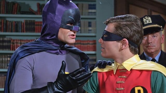 Opening shot: Medium close-up of Batman in purple cowl and cape with yellow bat emblem, facing Robin in red and green costume with yellow cape and black mask, a uniformed police officer in hat standing behind in a wood-paneled library with bookshelves.
Camera movement: Slow push-in from medium shot to close-up on Batman's face, steady and deliberate pacing to build tension.
Subject and action: Batman gestures emphatically with a gloved hand while speaking animatedly to Robin, who nods attentively, lips moving in response; police officer observes silently in the background.
Motion cues: Subtle parallax as foreground figures shift against static bookshelves, Batman's cape occludes part of Robin momentarily during gesture, depth evident in layered room elements.
Scene setting: Indoor library office with wooden shelves filled with books, daytime lighting from unseen windows casting soft shadows, serious and conspiratorial atmosphere.
Aesthetic and grade: Retro 1960s TV style with vibrant comic-book colors, warm saturated grade, slight film grain and soft focus emulating vintage broadcast footage; 50mm lens.