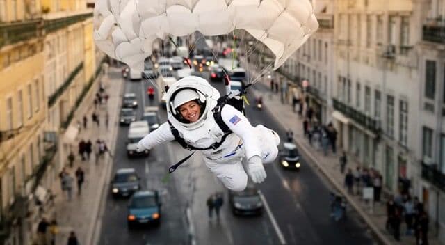 The camera pans downward as Pamela ejects and descends via parachute, landing with a thud on the street, sounds of wind rushing and impact echo, followed by surprised gasps from onlookers, suggested duration 8 seconds.