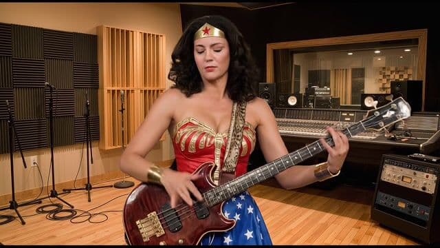 In a warmly lit recording studio with gray foam-paneled walls, wooden accents, and a mixing console in the midground, a Wonder Woman in her iconic red-and-gold corset, blue star-spangled skirt, golden bracelets, and tiara stands confidently holding her black Gibson bass guitar with gold hardware slung over her shoulder. She launches into a dynamic Stanley Clarke-inspired performance, her fingers slapping and popping the strings with fiery precision—right hand thumping the body for thunderous pops, left hand tapping harmonics and flying across the fretboard—while her body sways rhythmically, curly black hair bouncing and focused expression intensifying as deep resonant bass riffs fill the air amid faint cable hums and amp buzz. The 35mm lens camera steadily dollies in from medium-wide framing, foreground guitar neck blurring subtly faster than the static background panels and mic stands, under soft diffused overhead studio lights casting gentle shadows on the wooden floor, in hyper-realistic cinematic style with vibrant costume colors against neutral tones and immersive low-end bass groove audio.