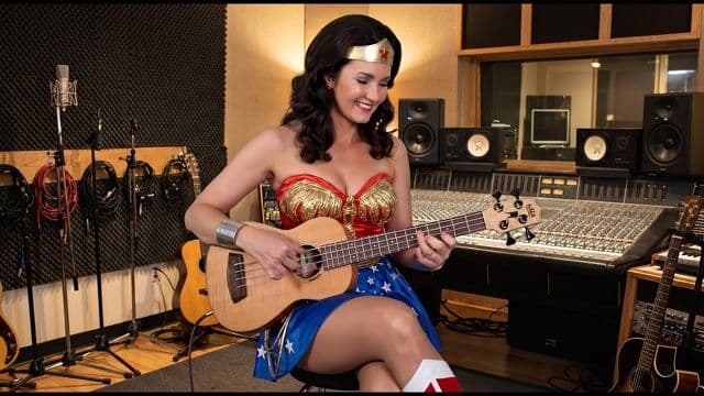 In a warmly lit recording studio with black foam-paneled walls, microphone stands, and a glowing mixing console in the depth of field, a curly-haired woman in a red corset, gold accents, blue star-spangled skirt, tiara, bracelets, and white starry boots sits on a stool, her fingers deftly plucking and strumming the yellow ukulele bass in intricate jazz fusion rhythms—starting with a steady groove that builds to lively riffs, her head nodding subtly and smile widening as she leans into the melody. The camera steadily dollies in from a 35mm lens at a measured pace, foreground strings vibrating sharply while background speakers and guitars remain parallax-still, enhancing depth. Golden studio lamps cast soft, diffused glows with subtle shadows on her skin and instrument, maintaining the vibrant color palette of reds, golds, and blues in a cinematic photographic style. Groovy jazz fusion bass lines fill the air with punchy plucks and reverb, accompanied by faint studio hum and her soft rhythmic breaths.