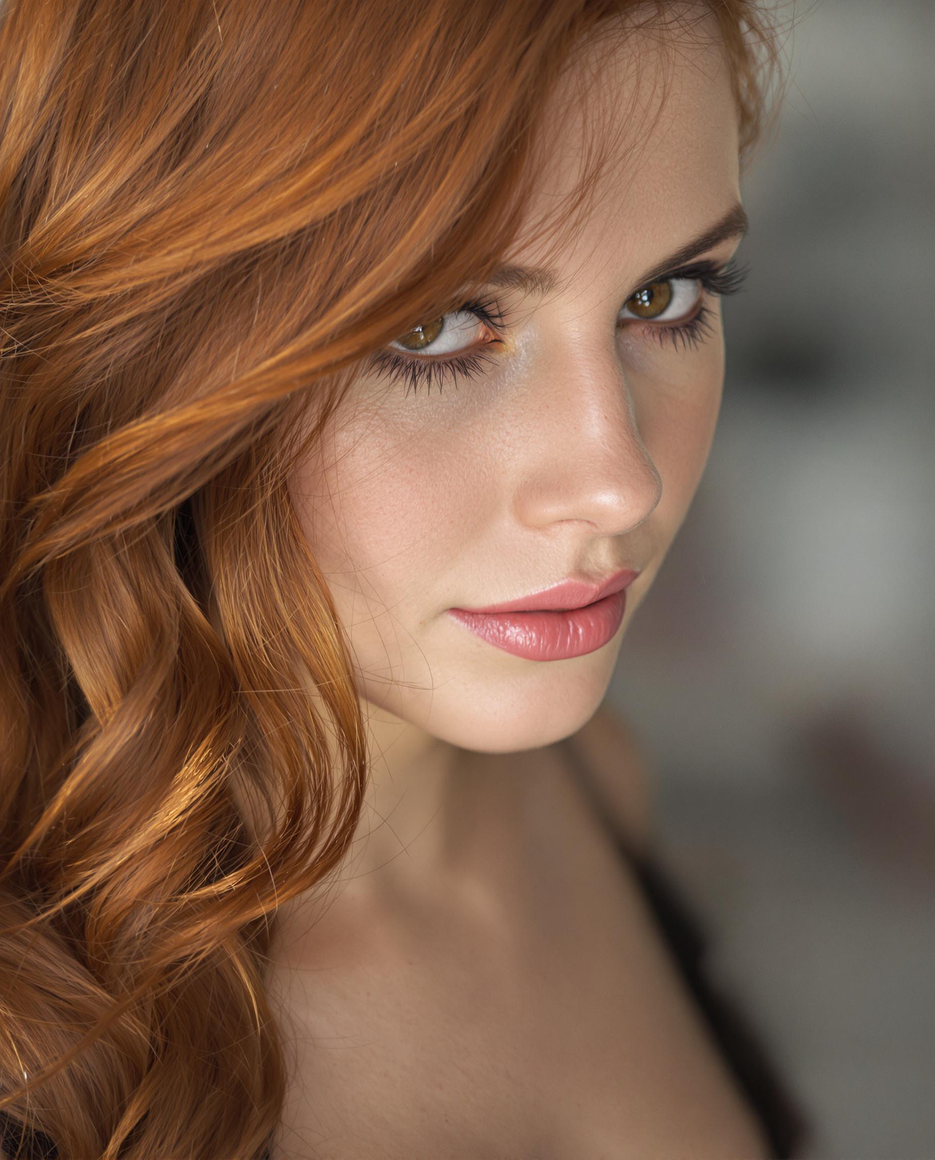 A highly detailed photorealistic portrait photograph of a young woman in her upper body, captured with a DSLR camera and 50mm lens for shallow depth of field, featuring soft cinematic lighting that imparts an ethereal glow to her smooth skin and rich auburn hair styled in a cascading side