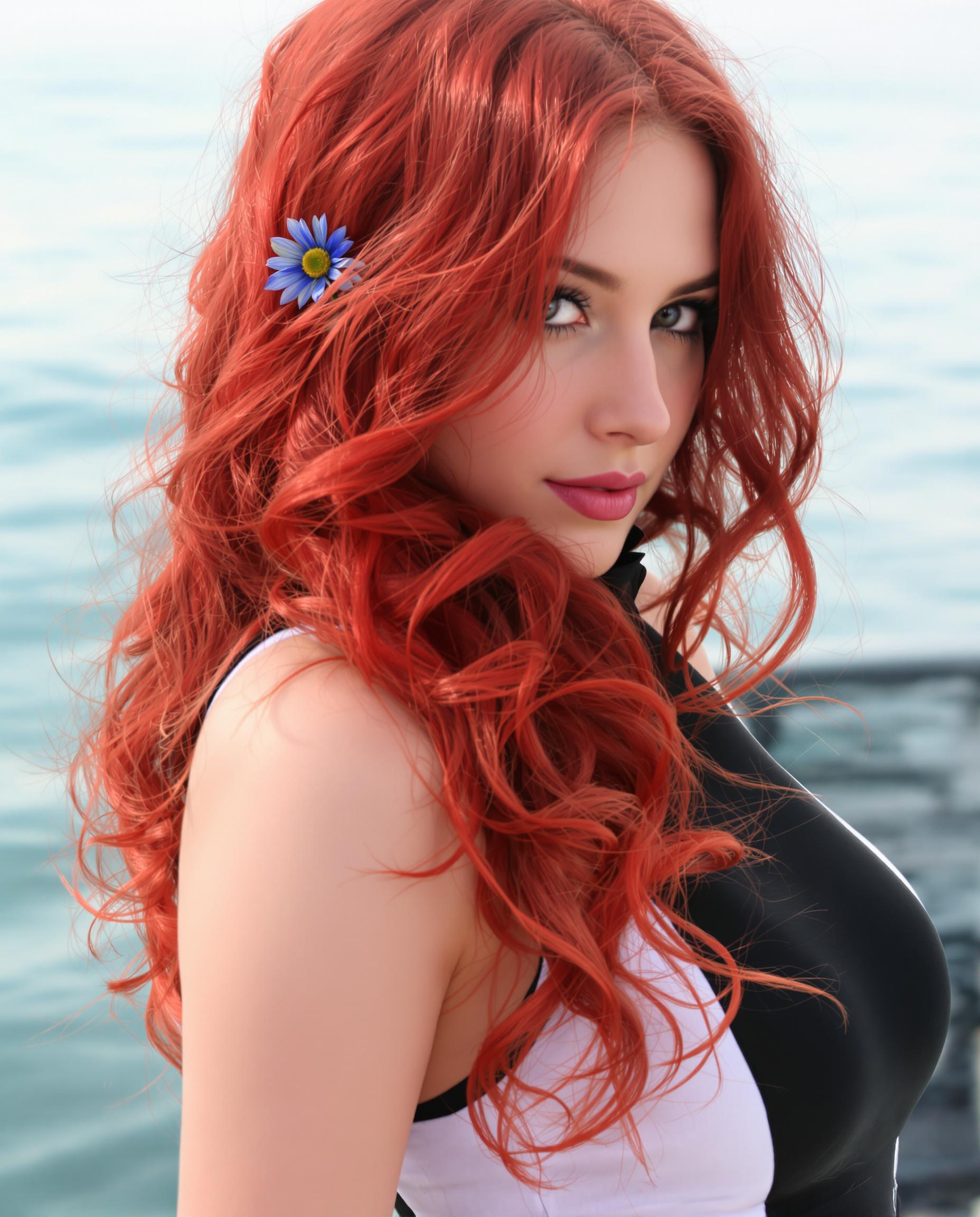 A photorealistic closeup portrait of a beautiful woman with long, flowing red hair cascading in detailed waves, subtle highlights and shadows adding realistic texture and volume, accented by a small blue flower tucked into the strands. She wears a fitted sleeveless top with a high neckline in black and white, featuring a visible back zipper and smooth, stretchy fabric with a slight sheen, set against a serene sea background under soft, diffused lighting that evokes a calm mood, rendered as a high-resolution digital painting with smooth color blending and intricate details.