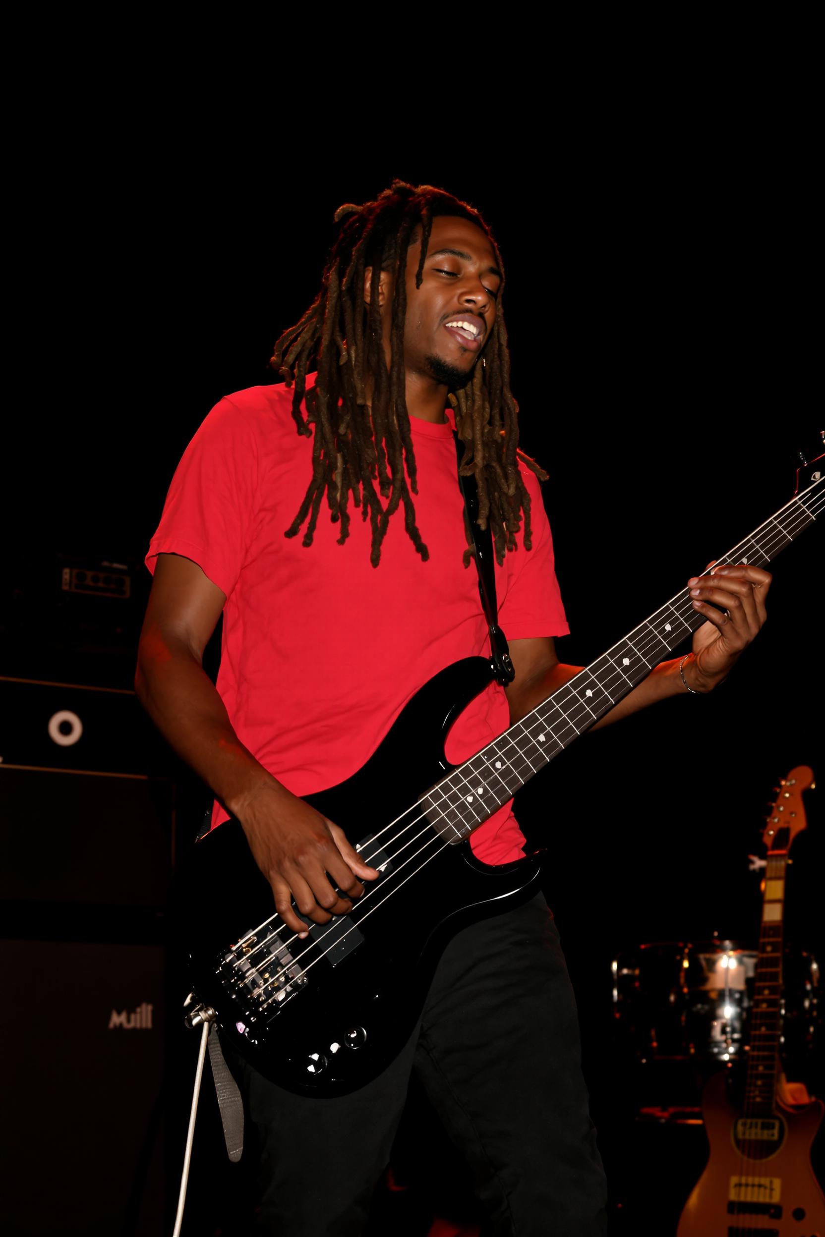 A highly detailed, realistic photograph of a young Black musician with long dreadlocks performing on a dimly lit stage, captured in a live concert setting with warm ambient lighting and deep shadows. He has a focused expression, mouth slightly open as if singing or concentrating, wearing a vibrant red short-sleeved t-shirt and black pants. He is playing a sleek black headless electric bass guitar with a strap, his left hand fretting the neck and right hand plucking the strings. In the background, blurred stage equipment including a black amplifier stack with circular logo, a silver drum set or pedalboard, and another guitar resting on a stand, all against a dark void-like backdrop. Photorealistic style, high-resolution digital photography medium, rich color palette dominated by reds, blacks, and warm yellow highlights from stage lights, emphasizing texture in hair, fabric, and glossy instrument surfaces, dynamic composition with slight motion blur on hands for energy.