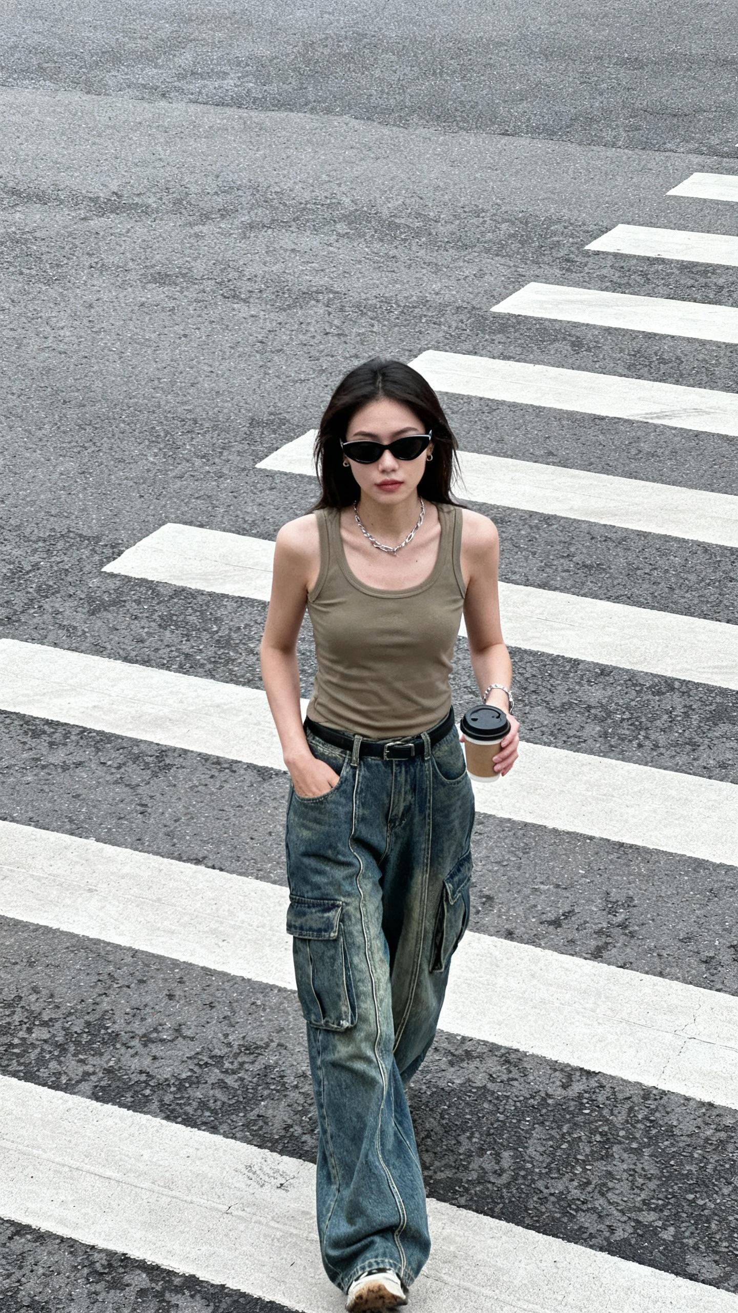 A young woman strides naturally across a broad pedestrian crosswalk, clad in baggy denim cargo pants detailed with visible seams and a khaki tank top neatly tucked in. She wears sleek black cat-eye sunglasses paired with understated silver jewelry, her relaxed pose marked by one hand slipped into a pocket and the other grasping a takeaway coffee cup. The setting is stark: just the textured asphalt beneath her feet and crisply painted, white zebra crossing lines stretching wide. Neutral daylight filters through soft overcast skies, casting gentle, diffuse shadows that lend a subdued atmosphere. Close attention reveals the textured weave of the denim, subtle wrinkles folding across the tank top, faint scuffs on her footwear, and porous skin illuminated with natural fidelity. Captured from a high vantage point, the framing is wide and candid, cropping part of her legs and blurring one arm slightly, conveying the authenticity of surveillance footage. The palette is muted and natural, the lighting and textures unembellished, embodying documentary-style realism in a moment frozen by an impersonal street camera.