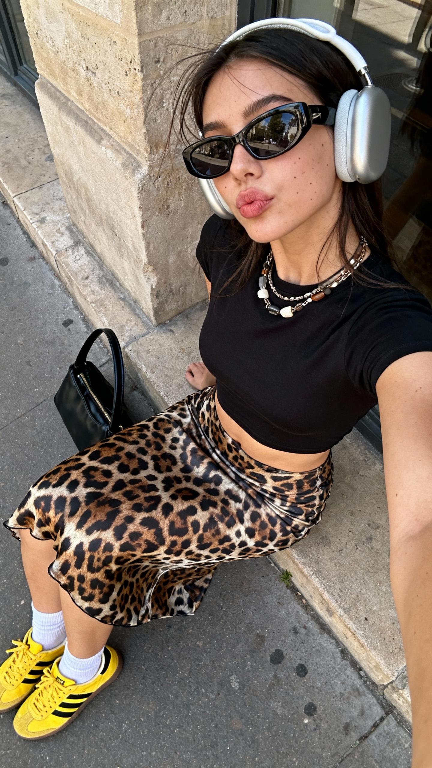 A candid, playfully spontaneous wide-angle iPhone selfie taken from a distinctly elevated overhead angle shows a young woman sitting casually on a city sidewalk ledge, leaning back slightly with her lips softly pursed, directly engaging the camera with a relaxed, neutral expression. She wears an original fitted and cropped black baby tee creatively reimagined without any prints, paired with a uniquely patterned slip skirt inspired by leopard motifs but distinctly stylized with inventive color and texture. Complementing the look are bright yellow sneakers featuring bold black stripes, casual white ankle socks, and an artfully placed black handbag resting on the ground nearby. Her accessories include large, modern headphones, oversized sunglasses with an original shape, and layered necklaces exhibiting varied textures and modern design elements. The authentic urban background features textured stone walls with subtle window reflections and natural daylight casting believable soft shadows and highlights. Textural realism highlights the fabric wrinkles of the tee and skirt, delicate hair strands partially visible under the headphones, natural skin textures with subtle imperfections, and detailed material surfaces of the handbag and sneakers. The composition emphasizes exaggerated wide-angle distortion by enlarging her upper body and face, capturing a spontaneous handheld selfie moment that reflects casual social media aesthetics, self-expression, and stylish urban authenticity.