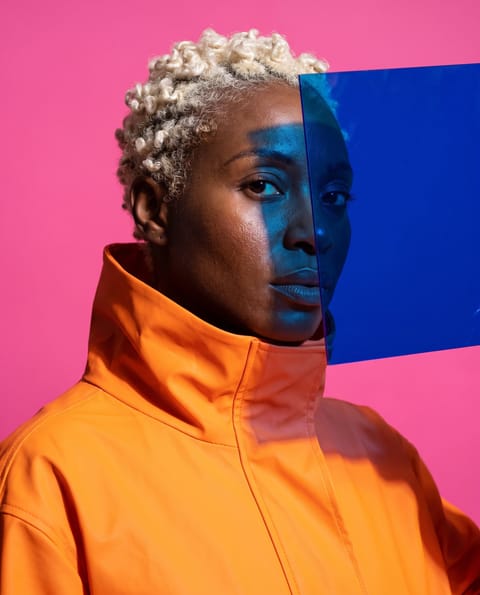 Editorial portrait of a woman with bleached curls in a tangerine high-collar coat, a cobalt acrylic sheet casting blue across half her face against hot pink