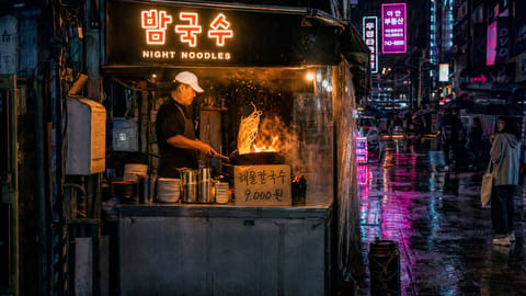 A cook tossing noodles over wok flames at a rainy Seoul alley stall at night under glowing hangul signs, pink neon on wet pavement