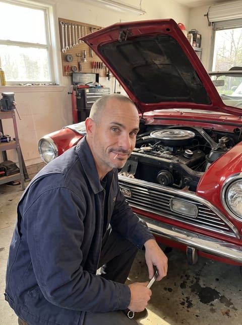 Brian crouched beside a red convertible with its hood open in a sunlit garage