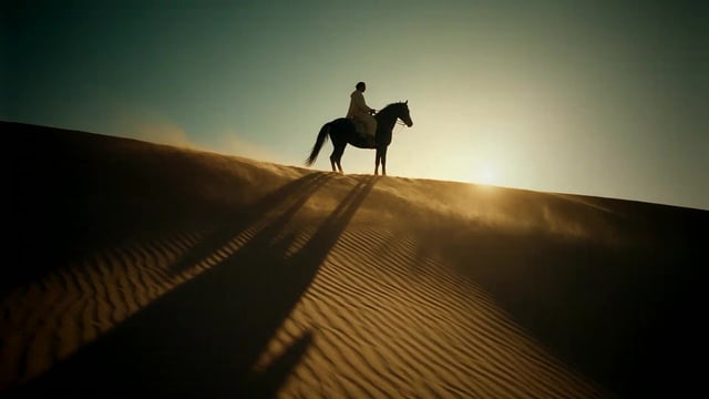 A lone rider on a black horse crests a sand dune at golden hour, dust catching the low sun