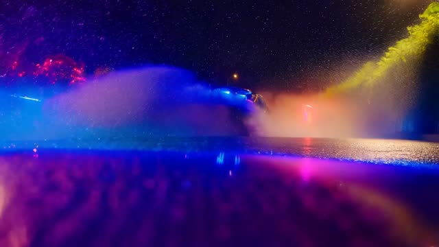 the ultra-wide-angle lens follows closely from the hood, with raindrops continuously splattering
against the lens. Ahead, a sports car speeds around a corner, its tires violently skidding against the wet road,
creating a mist of water. Neon lights refract in the rain, leaving colorful streaks on the car’s surface. The camera
swiftly shifts to the side of the car, capturing the wheels spinning at high speed, before finally moving to the rear