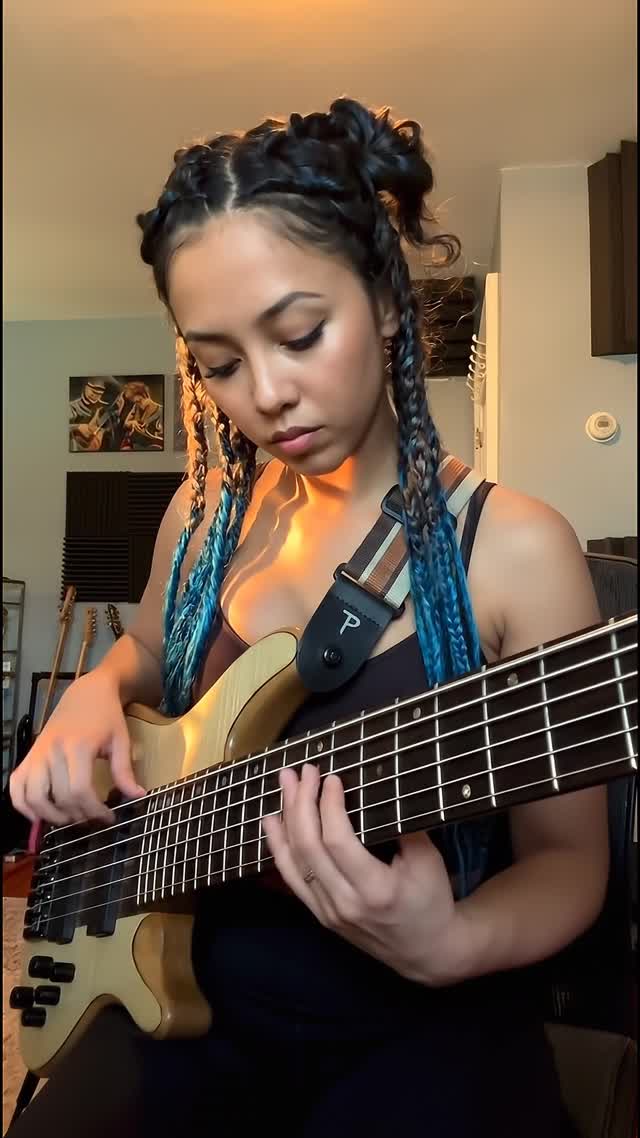 A focused young woman with blue-toned braided hair in double buns, wearing a black tank top and bass strap, stands in a warmly lit beige-walled room with golden sunset light streaming through window blinds, casting soft glows on her skin and cream-colored 5-string bass guitar with black pickguard. Her fingers begin to dance fluidly across the frets and strings in intricate jazz fusion style—slapping, popping, and plucking rapid chromatic runs and harmonics with precise intensity—while her body sways subtly to the groove, eyes locked downward in concentration, as the static bookshelf and wall art recede gently into depth. The 35mm lens camera steadily dollies in from medium shot to closer frame her expressive face and hands, enhancing foreground motion against the slower background parallax, under the persistent warm orange rim lighting and soft shadows. Cinematic photographic style with rich amber color grade and subtle grain; resonant bass lines thrum in jazz fusion rhythm, accompanied by faint room reverb and no other music.