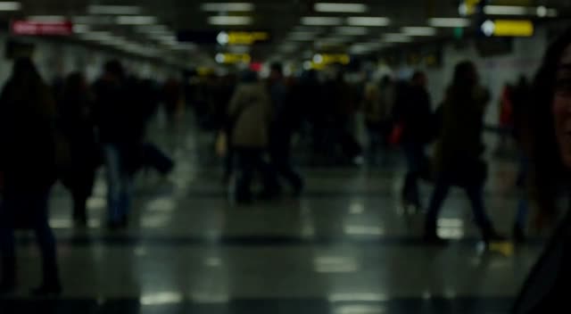In a dimly lit crowded subway station with flickering fluorescent lights and reflective tiled floors, a woman in a dark coat turns her body says "We're finally here, let's get this adventure started", and begins walking away from the camera at a steady pace. The camera tracks smoothly backward in a medium shot using a 35mm lens, following her through the throng of commuters who create shifting parallax layers in the foreground and background. Over eight seconds, she recedes deeper into the bustling scene, her figure gradually smaller amid the motion and atmospheric haze.