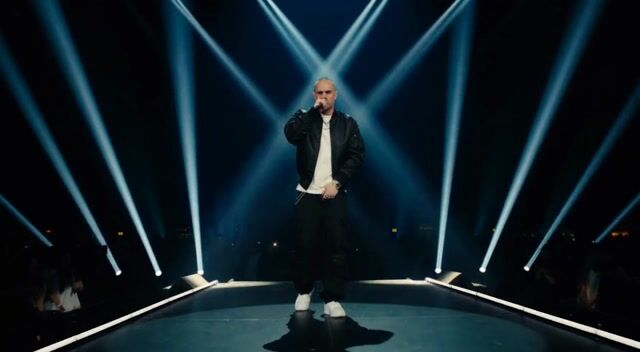 integrated_multimodal_description: Image1 defines the famous rapper's face, hairstyle, wardrobe, and exact appearance; preserve identity and clothing with zero drift. The rapper stands center stage under dramatic spotlights in a dark cinematic concert venue, mic in hand, delivering an intense performance. The camera executes a slow push in with small amplitude at slow speed from medium shot to close-up on his face and upper body as he sings with powerful energy and rhythmic gestures. <Rapper> (S1) says: <d>[English] I pull up clean, no cap new page, new plan, new stack PixelDojo.ai got the whole frame on lock Drag, drop, make it hit watch a rough idea click From blank screen to a flex I turn sparks into chips I build it fast, no stall</d> No on-screen text, no subtitles.
overall_soundscape: Live stage reverb on the rapper's powerful vocals and ad-libs from Audio1, faint crowd energy and microphone presence, subtle stage footsteps and clothing rustle.
non_diegetic_music: Sparse cinematic hip-hop beat with deep 808s and atmospheric synth pads sitting low under the vocals.
