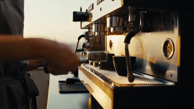 A barista in a narrow espresso bar tamps a portafilter, locks it into the group head, and starts the shot, at sunrise. Low warm sunlight comes through a window on the left, steam catches the light, brushed steel and matte ceramic surfaces. Begin on a medium shot of her hands at the machine, then slowly push in to a close-up of the espresso stream. Audio: the grind of the burr, the clack of the portafilter locking in, the hiss of the pump, and quiet morning room tone.