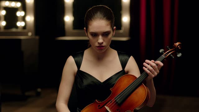 A young violinist sits alone backstage before a performance. Her fingers press hard against the neck of the violin and stop moving, her gaze fixes on the floor without blinking, her shoulders stay lifted, and she takes one short breath in and holds it. Medium close-up, static camera.