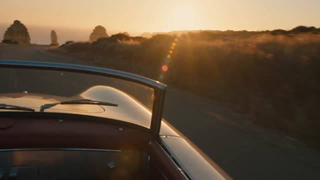Golden hour on a cliffside road. The camera tracks alongside a vintage convertible as it curves past dry grass and sea stacks, low sun flaring across the windshield. Warm amber grade, anamorphic bokeh, gentle handheld float. Audio: engine hum, wind, gulls.