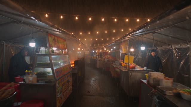 Handheld shot moving through a bustling night market in the rain. Strings of bulbs overhead, woks flaring, faces lit orange as the camera weaves between stalls. Saturated, energetic, slight lens spatter. Audio: sizzling, overlapping chatter, rain on canopies.