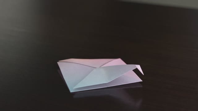 A 3D-animated origami crane unfolds from flat paper on a dark table, then takes flight, trailing a ribbon of soft light. The camera orbits as it rises. Clean studio lighting, pastel paper texture, Pixar-grade subsurface detail. Audio: paper rustle, a single airy swell.