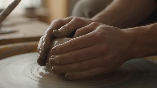 A potter's hands center wet clay on a spinning wheel, the wall of the vessel rising between thumb and fingers. Close, warm, tactile. Water beads and slips down the form. Soft north-window light, earthy palette. Audio: wheel hum, wet clay, breath.
