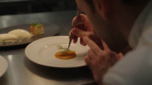 [0-6s] A chef plates a dish with tweezers under a warm pass light, tight over-the-shoulder. [6-12s] The camera pulls back to reveal the full kitchen mid-service, cooks crossing frame in choreographed rhythm. Documentary handheld, rich shadow. Audio: ticket printer, pans, terse callouts.