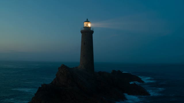 Slow orbit around a weathered stone lighthouse on a headland at blue hour. The beam sweeps toward camera once, catching sea spray in the air. Cool cyan sky against warm lamp glow. Audio: surf on rock, a low foghorn.