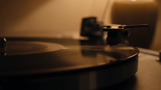 A vinyl record lowers onto a turntable in extreme close-up. The stylus settles into the groove, dust motes drifting in a warm lamp beam behind it. Shallow focus, nostalgic amber grade, 35mm texture. Audio: needle drop crackle, then soft room tone.