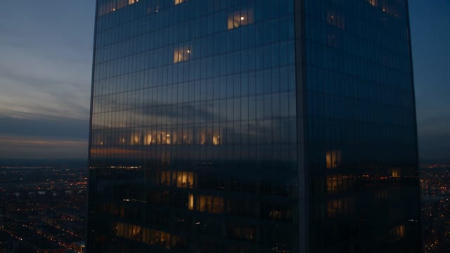 The camera cranes up the face of a glass tower at dusk, cloud reflections sliding across the panels, city lights blinking on floor by floor until it clears the roofline into open sky. Cool blue with warm window pinpricks. Audio: city hum falling away, wind at altitude.