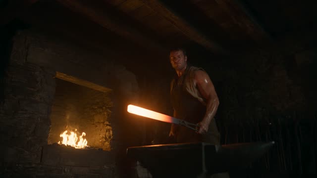 A blacksmith draws glowing steel from the forge and sets it on the anvil. Sparks burst on the first hammer blow and arc through a dark workshop. Firelight as the only key, deep shadow, high-speed clarity on the sparks. Audio: ring of hammer, roar of the fire.
