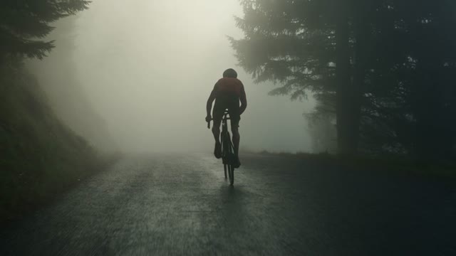 A cyclist crests a mountain pass into fog at dawn, breath and wheel spray catching the light, the camera tracking just ahead on a gimbal. Muted greens and greys with a single warm jersey accent. Audio: tyre buzz on wet tarmac, hard breathing, birdsong.