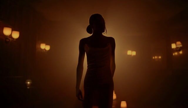 A confident woman in a silk evening dress walking slowly through a dim jazz lounge, warm lamplight, shallow depth of field, cinematic slow motion
