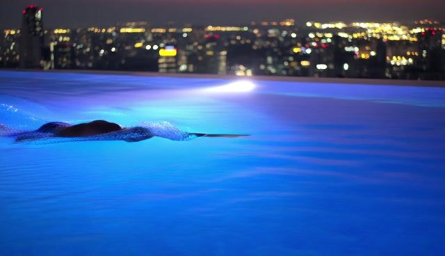 A fit man swimming a slow lap in a rooftop infinity pool at dusk, city lights bokeh, water rippling, cinematic