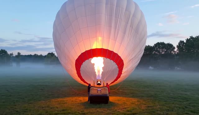 A hot air balloon inflates at dawn on a misty field, flame roaring as the envelope rises. One continuous take, slow pull back. Burner blasts and canvas rustle.