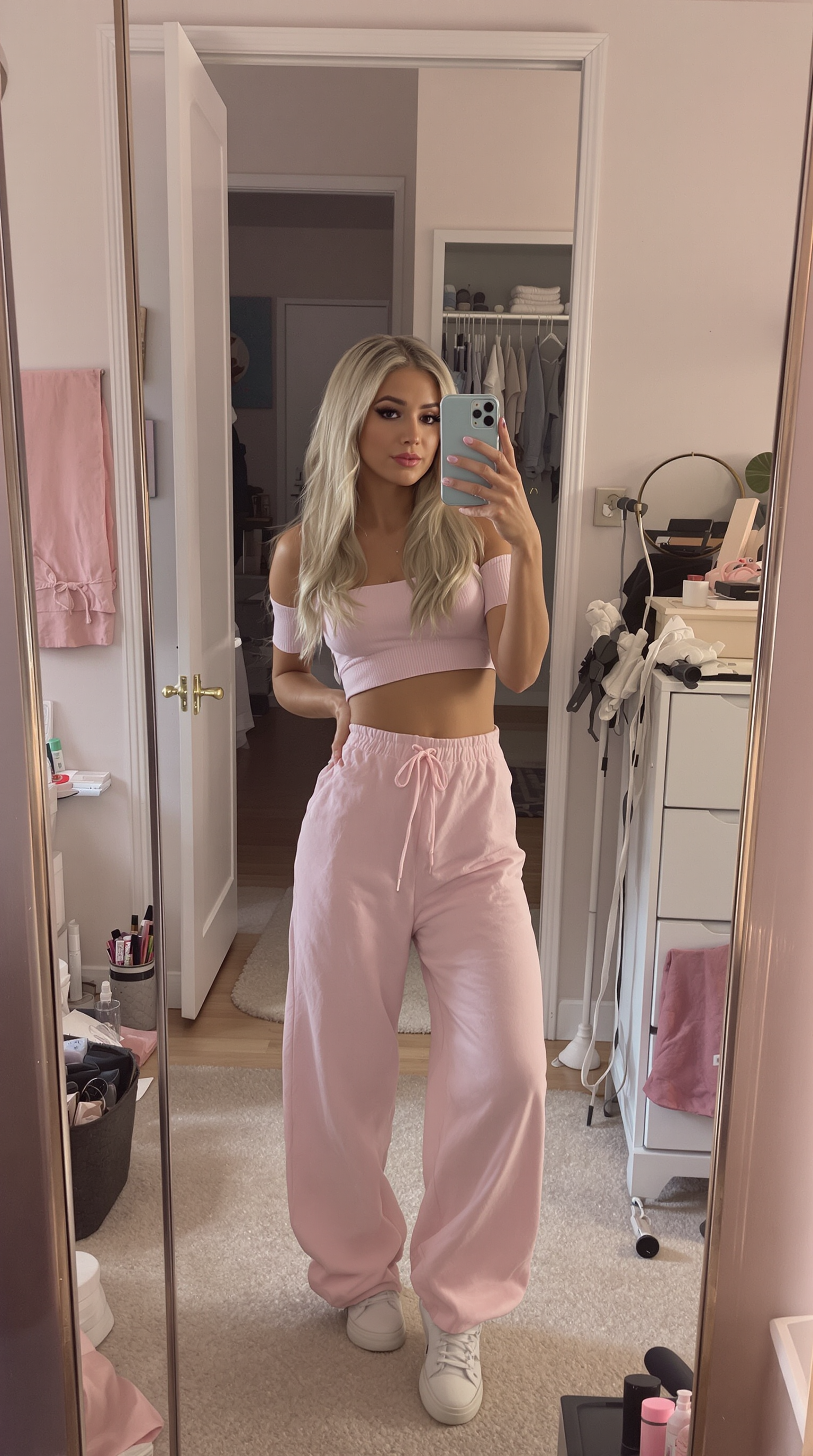 Stylish mirror selfie featuring a 25 years old woman with ash-blonde hair and long false lashes. She’s dressed in a casual crop top and lounge pants combo, posing effortlessly with a hand on her hip. The room has a modern aesthetic, with a large mirror, scattered makeup items, and soft lighting that enhances the pastel colors around her. The angle of the shot is slightly tilted, capturing a laid-back, influencer-inspired vibe