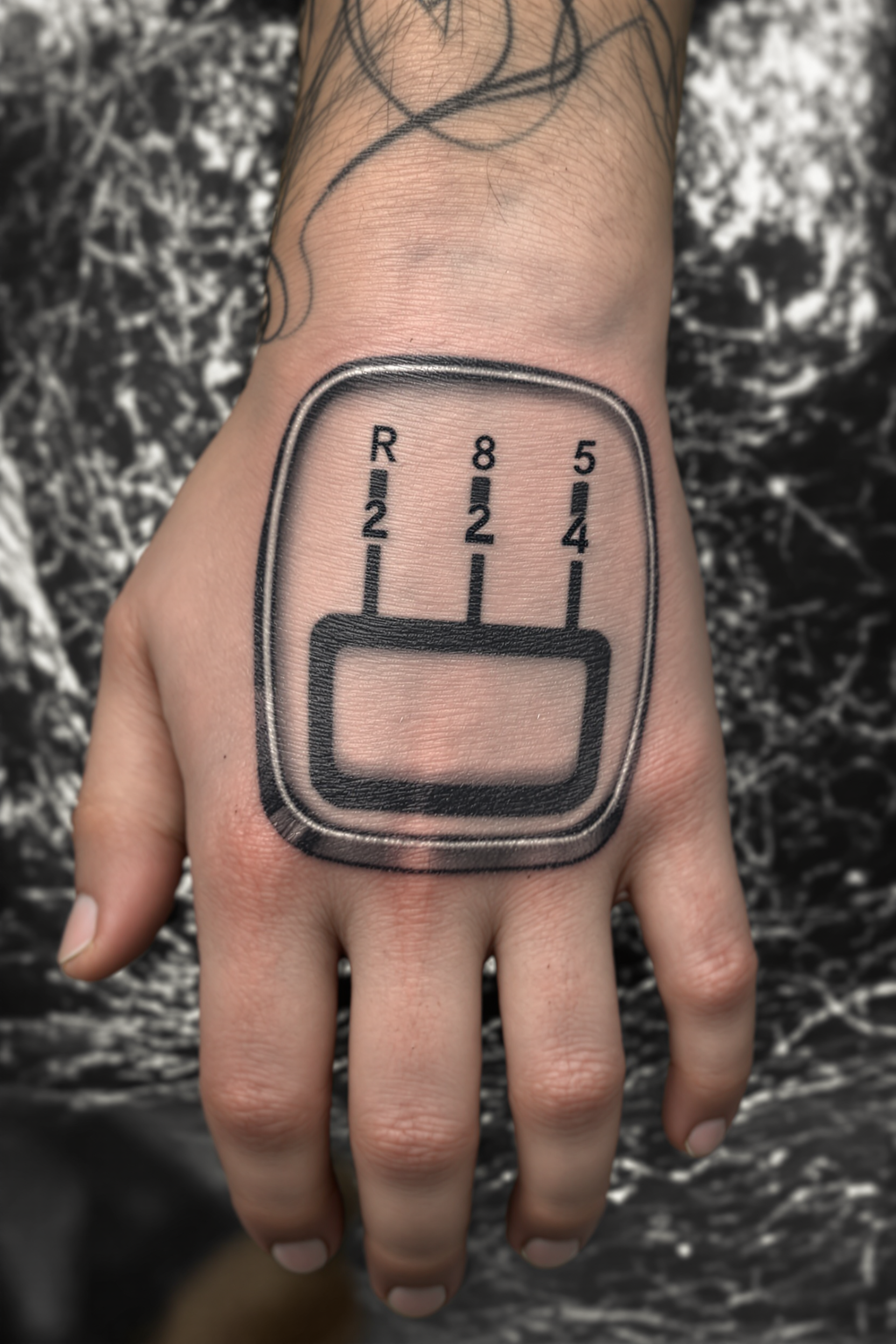 This image features a tattoo on a persons hand. The tattoo is a highly detailed and realistic representation of a gear shift lever, which is a common symbol for mechanics, drivers, or those with an affinity for cars and vehicles.The gear shift lever is depicted with a series of steps and notches, each labeled with a letter and number, indicating the different gears of a vehicles transmission. The steps are arranged in a cross pattern, with the top row labeled R Reverse, 2, 4, and the bottom row labeled 6, 8, 10, 5. The gear shift lever itself is encased in a metalliclooking housing, which is detailed with realistic textures and highlights that give it a threedimensional appearance.The art style of the tattoo is hyperrealistic, with a high level of attention to detail and shading that makes the tattoo appear as if it could be a photograph of a real gear shift lever. The colors used are primarily metallic grays and blacks, with some shading and highlights that give the illusion of depth and dimensionality.The medium used to create the tattoo is ink, which is applied to the skin with a tattoo machine. The ink is carefully placed to create the illusion of the gear shift lever and its housing, with the artist skillfully blending colors and shading to achieve the realistic effect.Overall, the tattoo is a striking and detailed representation of a gear shift lever, designed to stand out and be easily recognizable. The hyperrealistic style and attention to detail make it a visually compelling piece of body art.